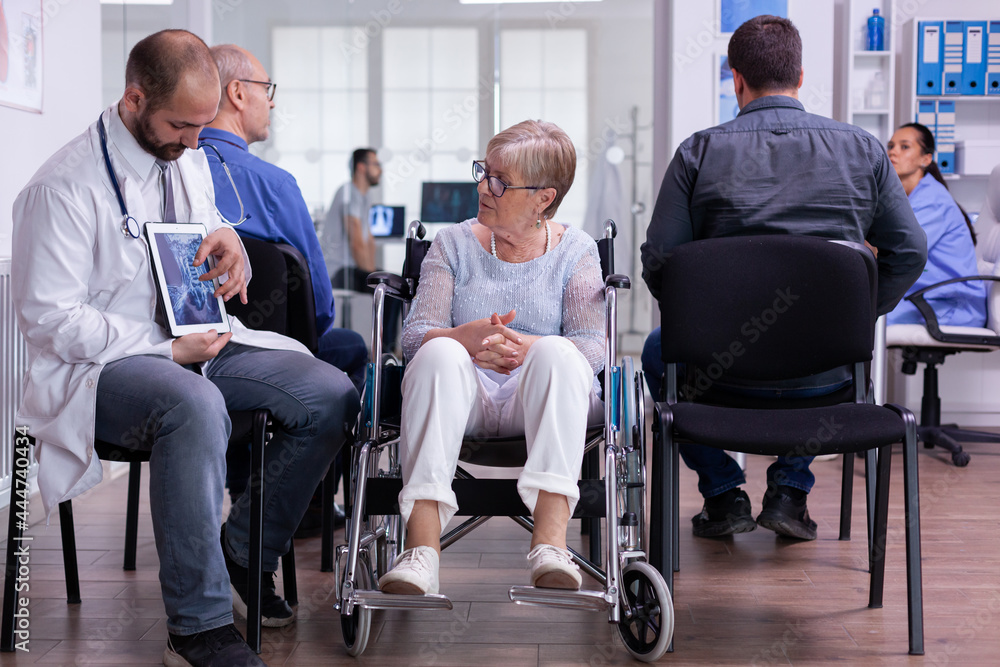 Doctor man sitting in waiting hospital area with senior disability woman discussing recovery treatment against middle aged illness. Doctor showing bones radiography using tablet computer