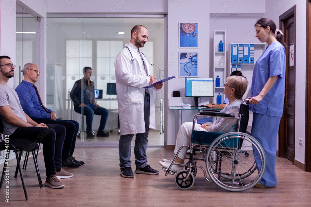 Doctor checking diagnosis of paralyzed, disabled senior woman in wheelchair sitting in reception room of hospital. Medical staff taking care of invalid elderly patient, man waiting in examination room