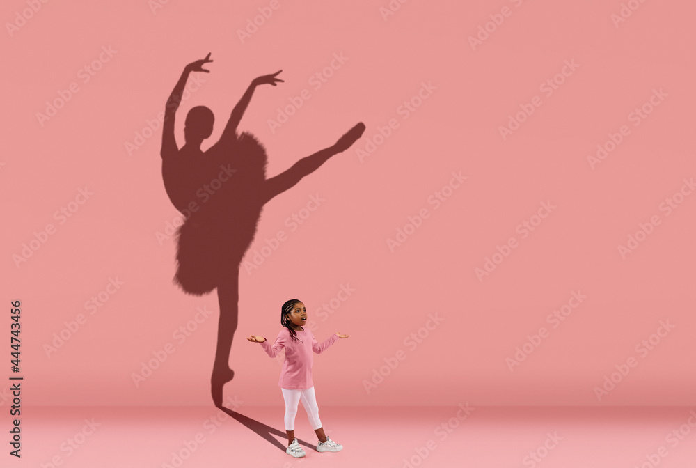 Childhood and dream about big and famous future. Conceptual image with girl and shadow of female ballet dancer on coral pink wall, background.
