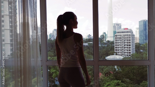 Young woman uncover the big window and looking out her apartment on the city buildings