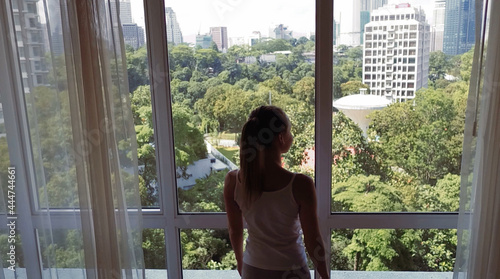 Young woman uncover the big window and looking out her apartment on the city buildings