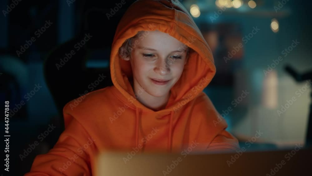 Cute Young Boy in a Hoodie Using Laptop Computer in Cozy Dark Room at Home. Happy Teenager Browsing Educational Research Online, Chatting with Friends, Studying School Homework. Cute Young Boy in a Hoodie Using Laptop Computer in Cozy Dark Room at Home. Happy Teenager Browsing Educational Research Online, Chatting with Friends, Studying School Homework.