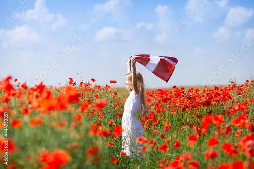 Blond girl holding flag of Latvia in the poppy field. Declaration of Independence Day. Ligo. Proclamation of the Republic of Latvia. Travel and learn latvian language concept. photo