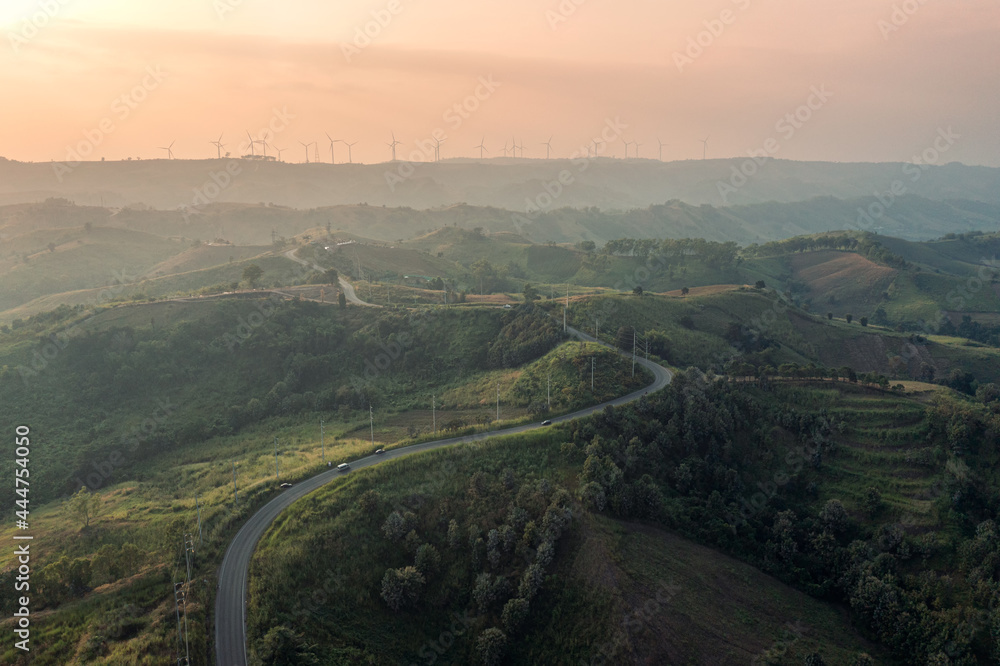 Aerial view of Curved highway on green hill and wind turbine on peak at sunset in countryside