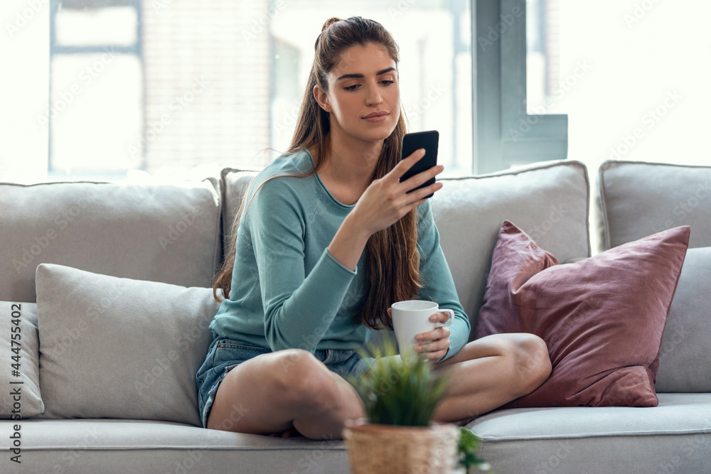 Pretty young smiling woman using mobile phone while drinking cup of coffee sitting on a couch at home.
