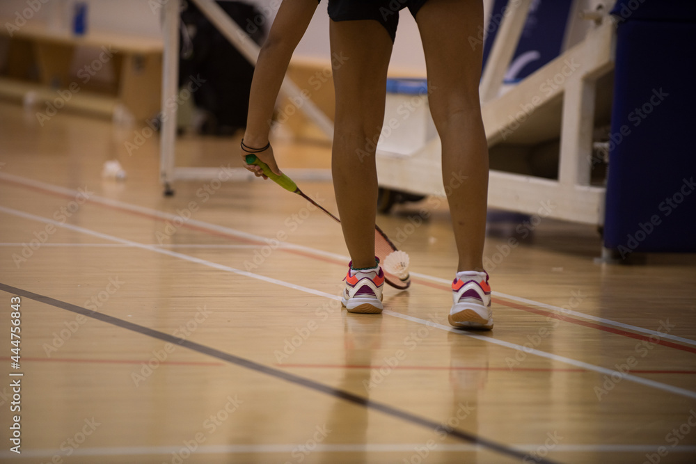 Female badminton player picks up a shuttlecock placed on wooden court while playing a match in a tournament. Female badminton player picks up a shuttlecock placed on wooden court while playing a match in a tournament.