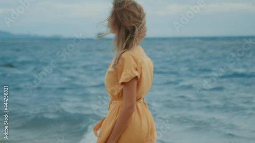 A slim blonde in a yellow dress walks along the beach and looks around