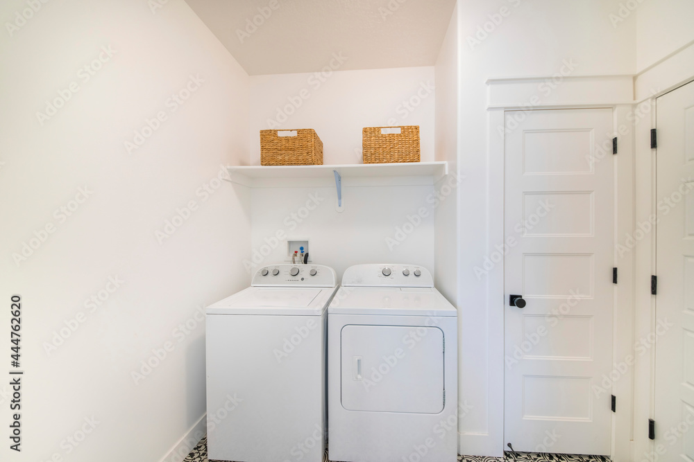 Small white laundry room interior with washer and dryer unit beside a narrow white door - obrazy, fototapety, plakaty 