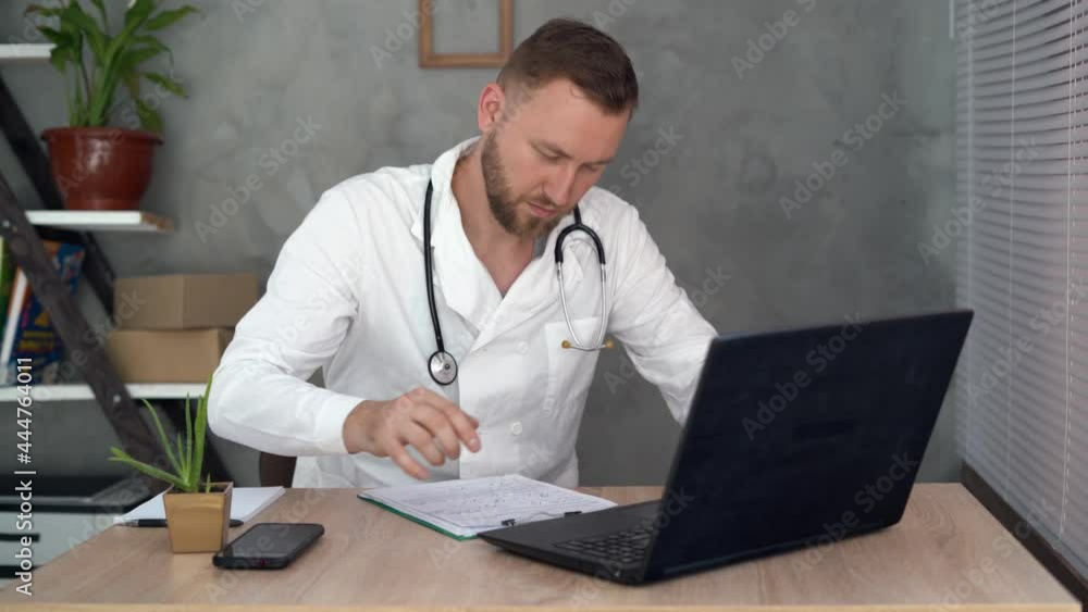 A young male doctor in a white medical coat sits in the office at the table and enters data from the table into his laptop. A young male doctor in a white medical coat sits in the office at the table and enters data from the table into his laptop.