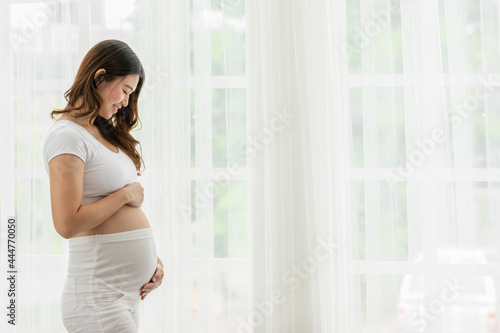 Happy Pregnant Woman standing in front of windows and stroking her big belly with love at cozy home,Pregnancy of young woman enjoy with future life relax with love,Motherhood and Pregnant Concept