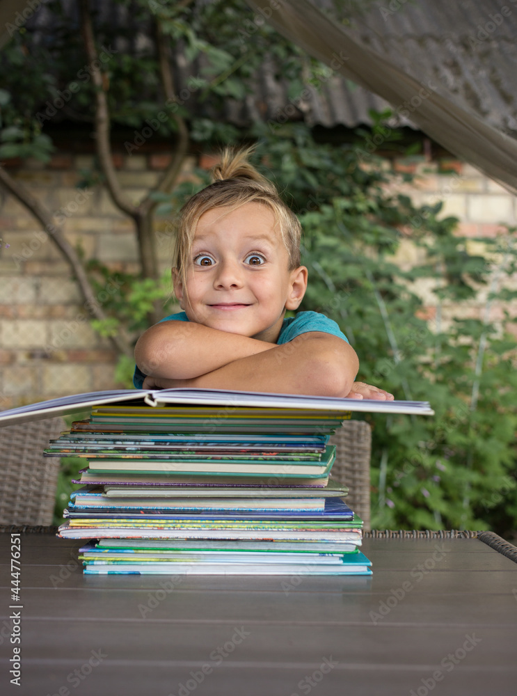 funny inquisitive preschooler sits at a table in the yard, in front of him is a large pile of children's books. Reading books, children's fantasies, interesting childhood. instill a love of literature