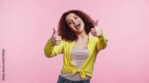 excited young woman showing thumbs up isolated on pink