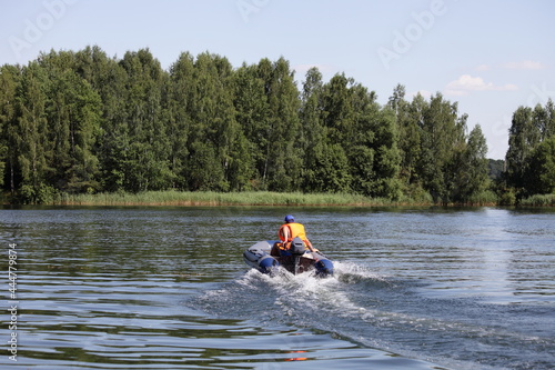 One man in orange lifejacket on inflatable motor boat with outboard motor fast floating on the river on green trees background , active recreation on the water at Sunnny summer day