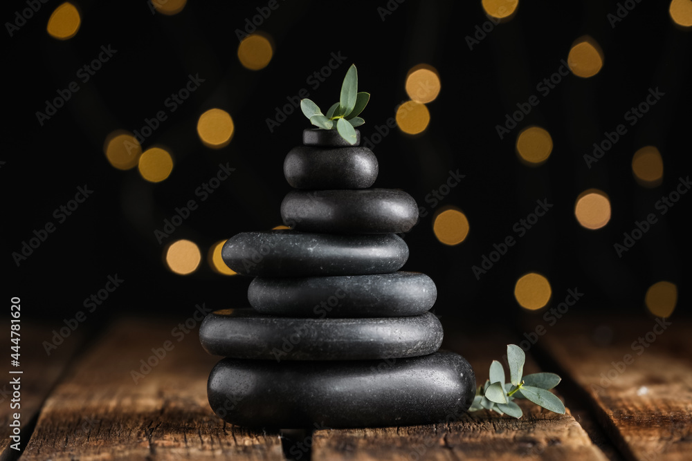 Obraz premium Stack of black spa stones with green branches on wooden table against blurred lights