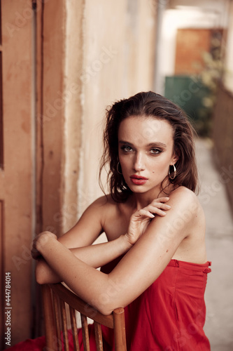 Charismatic brunette in a long red dress with beautiful facial features. sitting on a chair on the balcony in a small Spanish street. Painted red lips and incredibly beautiful brown eyes