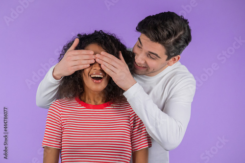 White man closes eyes of his beloved african american girlfriend before surprise her. interracial couple on purple studio background. Love, holiday, happiness concept.