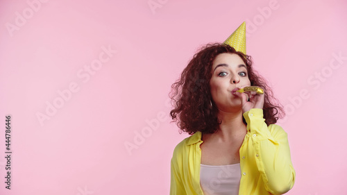 curly young woman blowing party horn isolated on pink