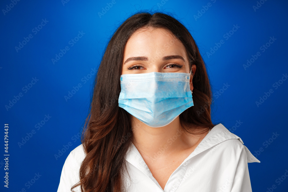 Young beautiful brunette woman wearing medical face mask against blue background Young beautiful brunette woman wearing medical face mask against blue background