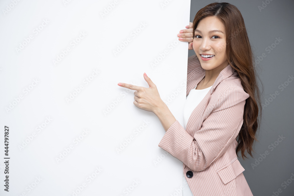 Cheerful Young Asian woman is standing behind the white blank banner and pointing to empty copy space advertisement board on grey background, Looking at camera Cheerful Young Asian woman is standing behind the white blank banner and pointing to empty copy space advertisement board on grey background, Looking at camera