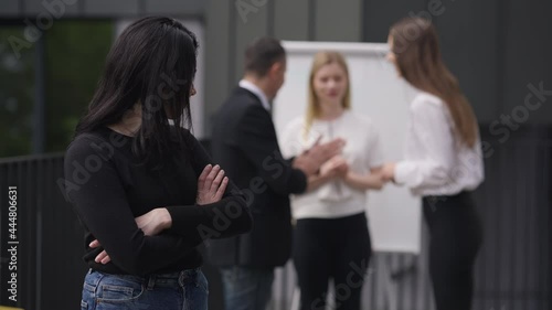 Shy Caucasian woman crossing hands looking back at colleagues discussing business project. Portrait of hesitant unconfident new employee at meeting outdoors. Slow motion