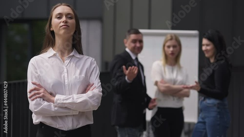 Portrait of confident arrogant woman crossing hands looking at camera ignoring colleagues mocking at background. Young beautiful Caucasian employee posing outdoors in slow motion