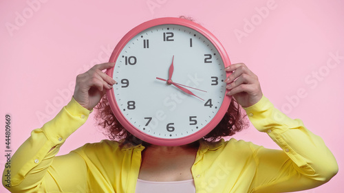 young woman covering face while holding clock isolated on pink