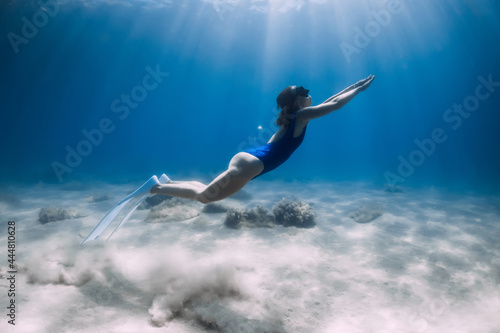 Lady freediver with white fins posing and glides underwater in blue ocean with sunlight.
