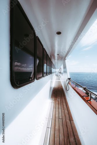 A wide-angle vertical shot of the side-deck of a luxurious white safari boat with a row of windows of the deckhouse wooden floor of the ways and the waterscape astern with a blue sea water © skyNext