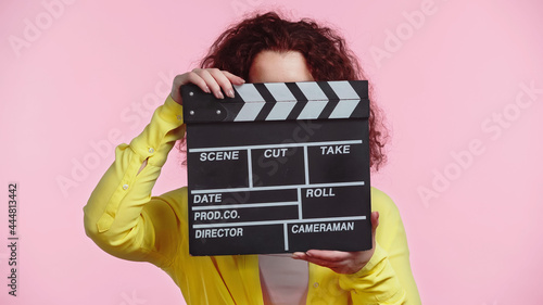 curly woman obscuring face with clapboard isolated on pink photo