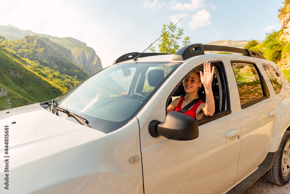happy young caucasian girl driving white off-road car down the mountain and waving. travel and adventure concept.