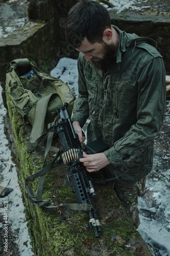 Portrait of airsoft player in professional equipment loads a gun with bullets in the forest. Soldier with weapons at war