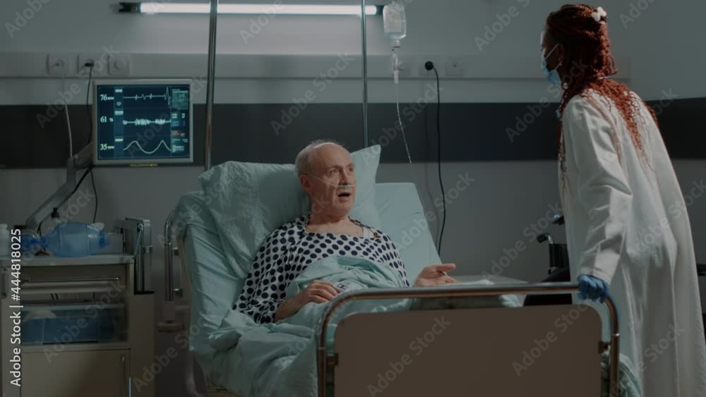 African american medic fixing adjustable bed for patient in hospital ward at medical clinic. Unwell old man with nasal oxygen tube looking at modern tablet for examination results African american medic fixing adjustable bed for patient in hospital ward at medical clinic. Unwell old man with nasal oxygen tube looking at modern tablet for examination results