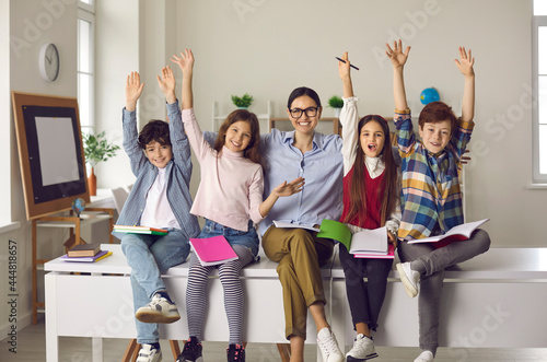 Education, successful completion of school year. Happy smiling teacher hugging overjoyed excited elementary pupils diverse group sitting on desk with raised hands up. Modern classroom interior