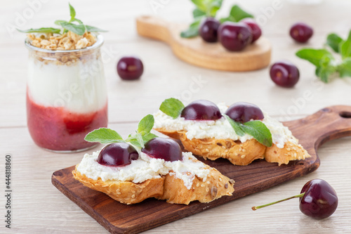 Sweet toasts with cream cheese, fresh ripe cherry and mint leaves decor on wooden board and miniature glass jar of aquafaba dessert with fruit jam and granola. Vegetarian snacks. Gluten free toasts.