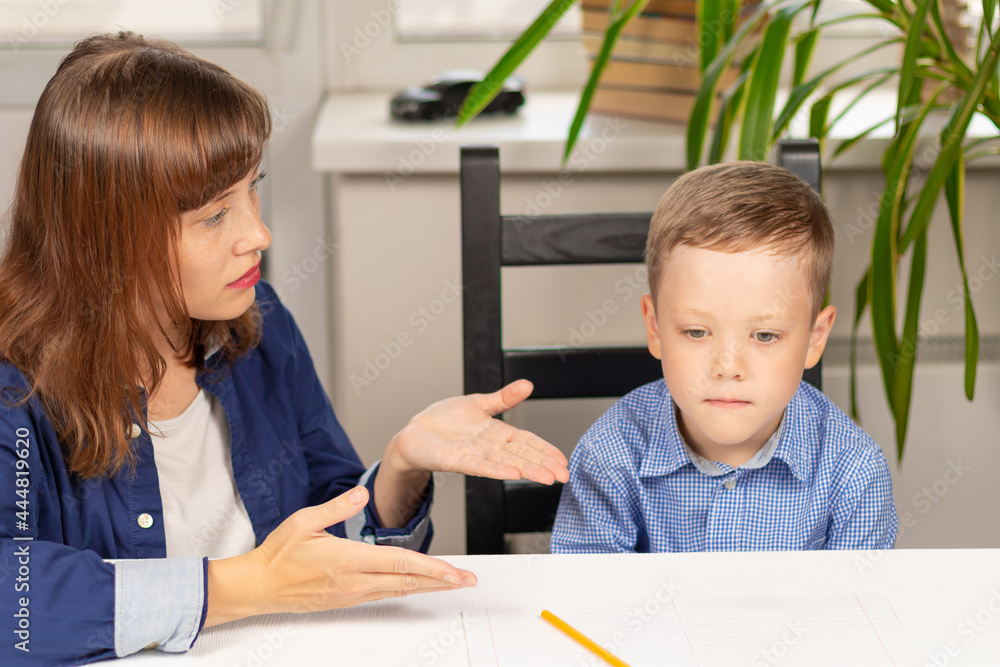Cute seven year old baby boy doing homework at home with mom on a white wooden table during a pandemic. Selective focus. Close-up. Portrait Cute seven year old baby boy doing homework at home with mom on a white wooden table during a pandemic. Selective focus. Close-up. Portrait