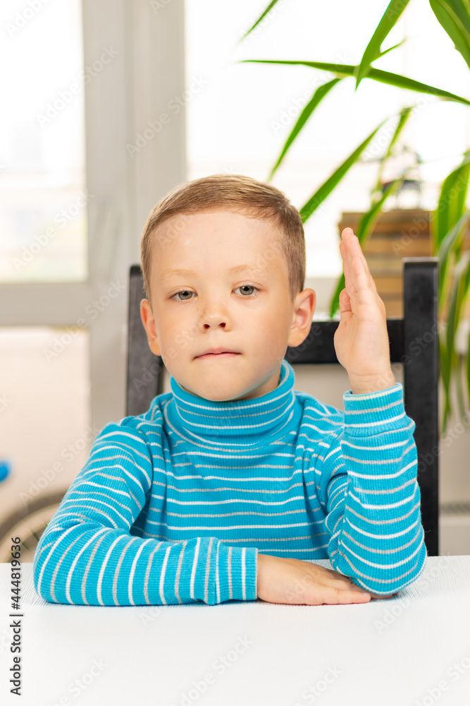 A cute seven-year-old child boy sits at home at a white wooden table against the background of a light window. Selective focus. Close-up. Portrait A cute seven-year-old child boy sits at home at a white wooden table against the background of a light window. Selective focus. Close-up. Portrait