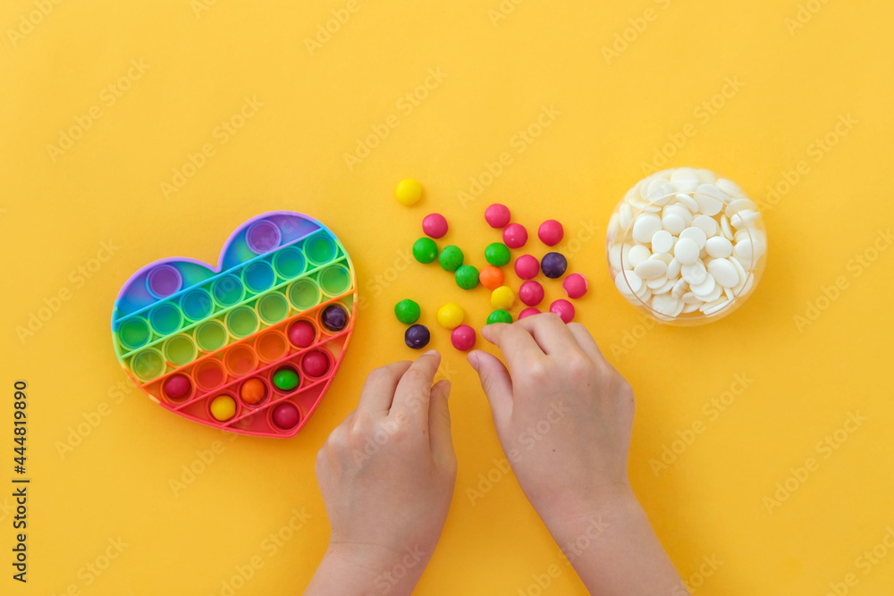 Use the popular pop it toy as a chocolate mold. Children is hands put colorful candies in a popit heart-shaped silicone mold on a yellow background. Step-by-step photo instructions. Step 2. Use the popular pop it toy as a chocolate mold. Children is hands put colorful candies in a popit heart-shaped silicone mold on a yellow background. Step-by-step photo instructions. Step 2.