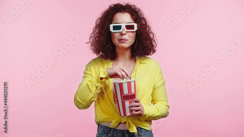 curly young woman in 3d glasses holding popcorn bucket isolated on pink