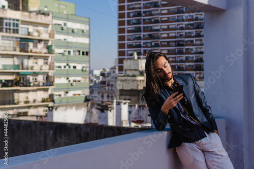 young hispanic latino male long haired businessman leaning on his terrace checking his smart phone. elegant, copy space