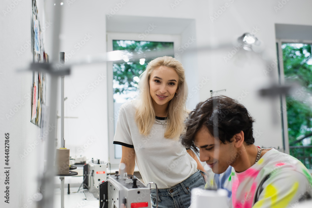 smiling designer looking at camera near tailor working on blurred foreground smiling designer looking at camera near tailor working on blurred foreground