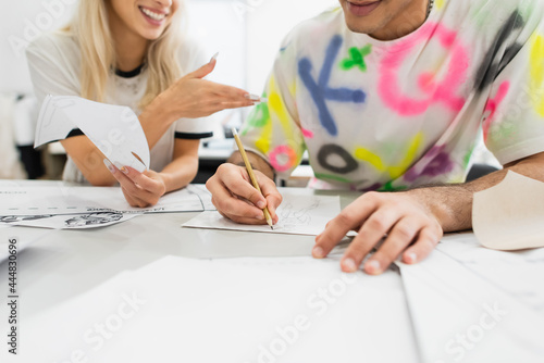 partial view of fashion designer drawing near smiling colleague pointing with hand, blurred foreground