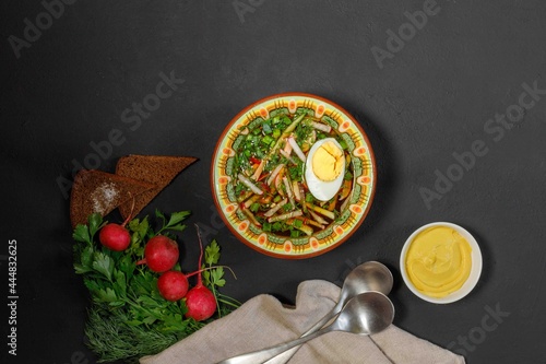 Cold soup with ham, egg, herbs. In a vintage painted bowl, on a black background with radishes, mustard and bread.Okroshka