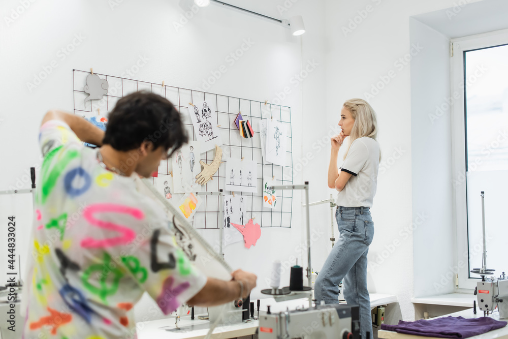 blonde fashion designer thinking near sketches on wall and blurred colleague with measuring tape blonde fashion designer thinking near sketches on wall and blurred colleague with measuring tape