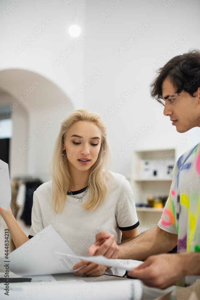 young fashion designer pointing at paper near blonde colleague in atelier