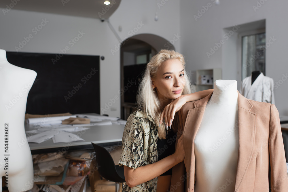 blonde fashion designer looking at camera while leaning on mannequin in atelier blonde fashion designer looking at camera while leaning on mannequin in atelier