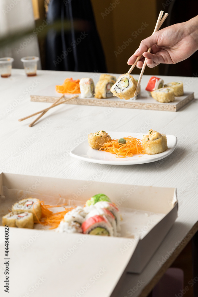 hand of a person organizing with chopsticks some sushi roll in a cardboard box and plates on a table in a restaurant, details of food texture, studio and lifestyle hand of a person organizing with chopsticks some sushi roll in a cardboard box and plates on a table in a restaurant, details of food texture, studio and lifestyle