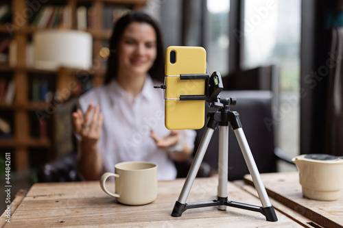 Happy young Caucasian millennial or gen z woman with long brunette hair streaming with smart phone on tripod, shooting social media blog in modern cafe. Influencer using social networks indoor. photo