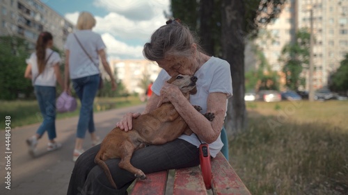 Senior 90-year-old woman with gray hair and deep wrinkles sits outdoors In assisted living facility on bench with small dachshund dog. Old female hugs and cuddles pet in the park on a bench photo