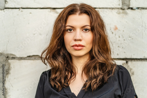 Front view portrait of adult caucasian woman posing in front of white wall outdoor in day looking to the camera wearing black dress with copy space and brown hair © Miljan Živković