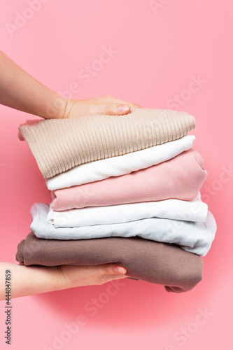 Female hands hold stack of knitted clothes on pink background close up.